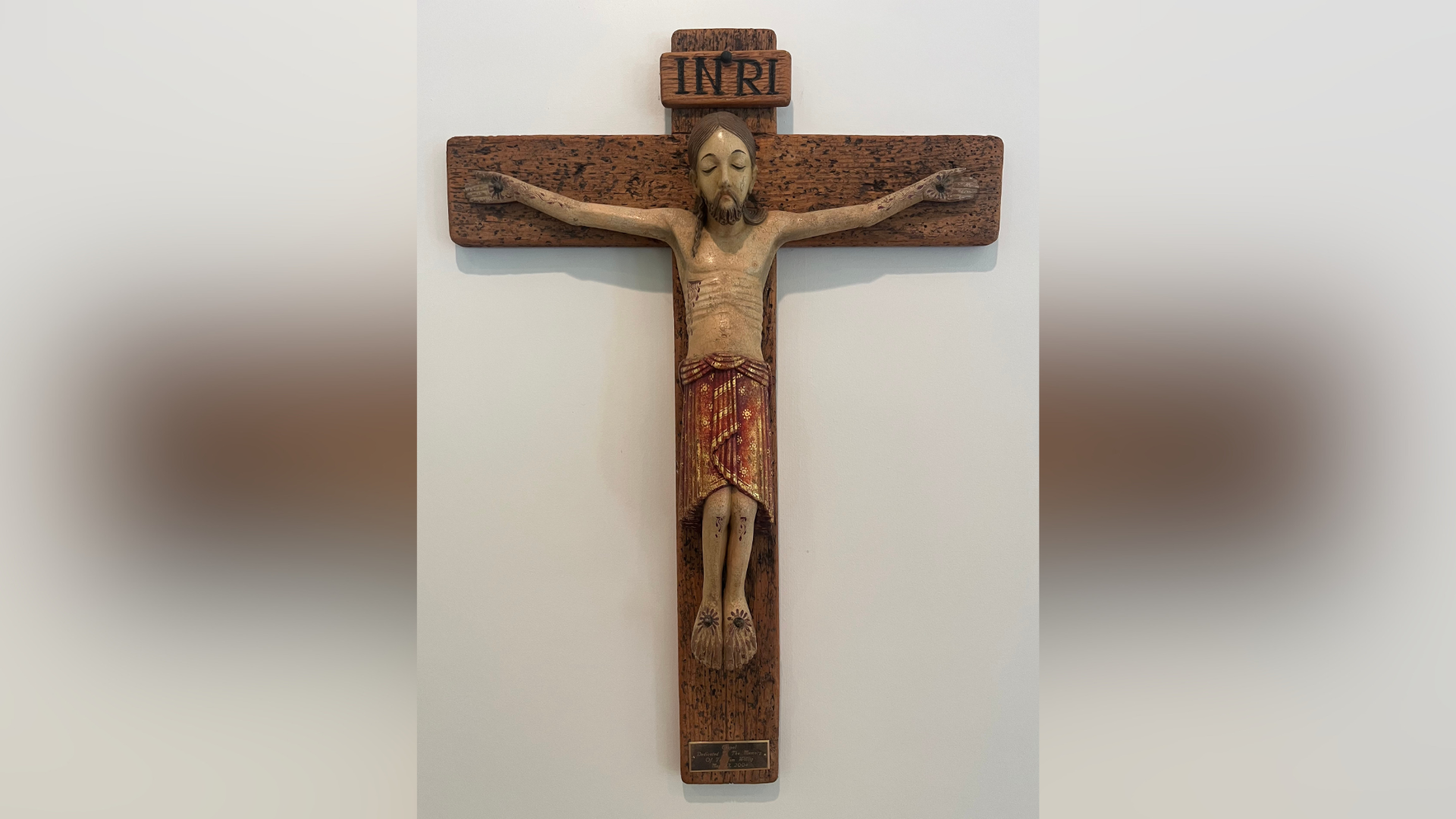A painted wooden crucifix mounted on a wall, featuring a polychrome carved figure of Christ with a red and gold loincloth, the INRI inscription on a titulus above, and a small dedicatory plaque at the base.